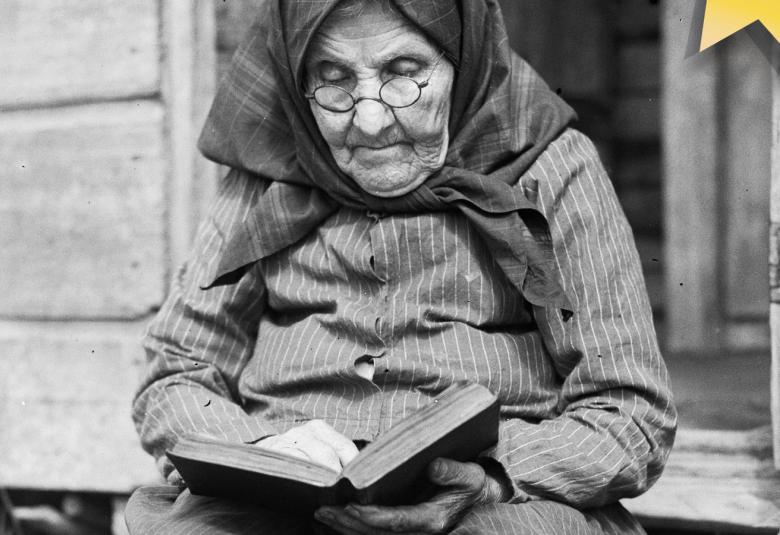 Kuvassa on vanha, ryppyinen nainen musta huivi päässään lukemassa kirjaa. Hän istuu portailla. Kuva on musta-valkoinen.