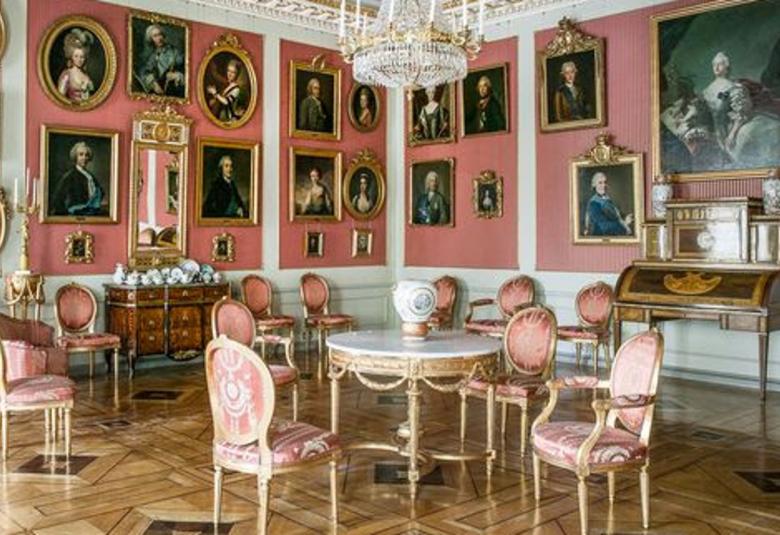 Kuvassa on etualalla kustavilaistyyliset valko-punaiset huonekalut ja vaaleanpunaisilla seinillä runsaasti pienikokoisia tauluja.  Katossa on on reunoilla koristeelliset ornamentit.