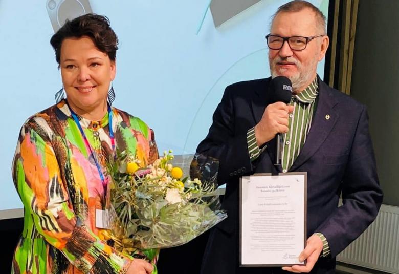 Kuvassa ovat Minna Kurttila värikkäässä mekossa ja kukat kädessään ja Erkki Kaila kunniakirja kädessään.  