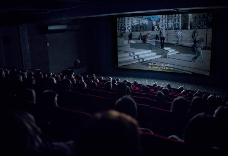 Kuvassa on pimeä elokuvasali, josta näkyy juuri ja juuri yleisöä.  Valkokankaalla näkyy näytettävä elokuva.
