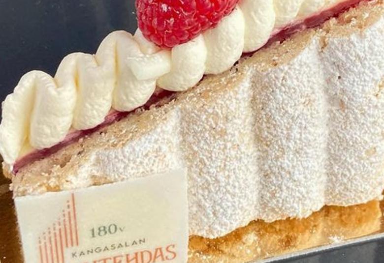 Kuvassa on vaalean ruskea urkujen muotoinen leivos, joka on nouseva ja päällä on valkoinen kermavaahtopursotus ja keskellä on punainen vadelma. Vasemmalla alhaalla on laatta, jossa lukee 180 vuotta Kangasalan urkutehdas.