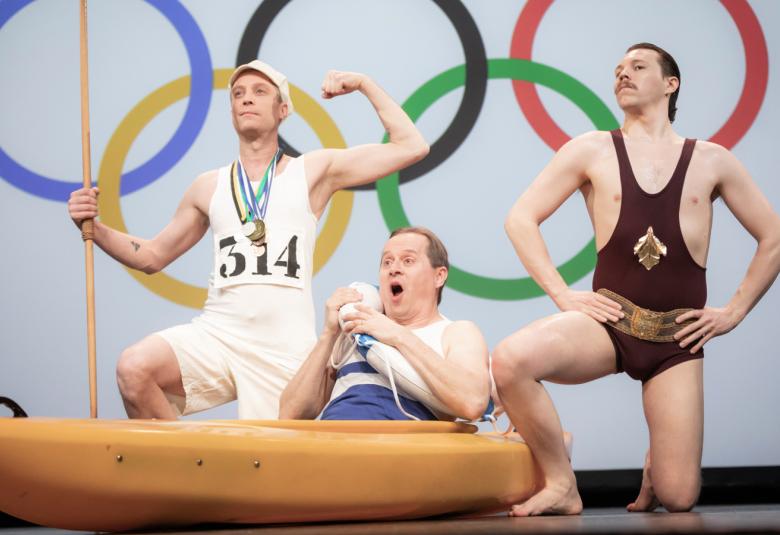 Kuvan tausta on valkoinen ja siinä on isot olympiarenkaat. Edessä on kolme miestä vanhanaikaisissa voimisteluvaatteissa ja he pullistelevat lihaksiaan.