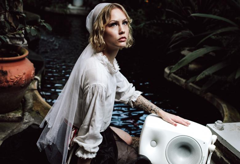 Kuvassa on Vilma Jää istumassa valkoisessa puserossa ja hänen huivistaan lähtee tyllilaahus.  Taustalla on ruskea ruukku, jossa on viherkasvi ja hän nojaa valkoiseen kaiuttimeen.