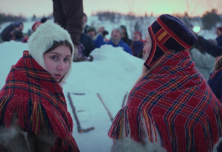 Kuvassa on kaksi nuorta naista Lapin puvuissa ja puna-valkoruutuisissa isoissa huiveissa. Takana näkyy lunta ja kauempana muita ihmisiä.