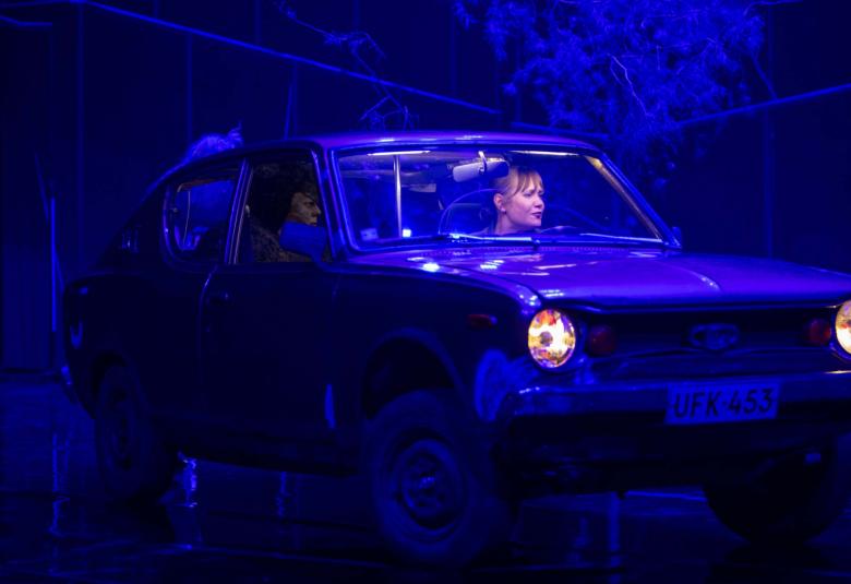 Kuvassa on Ella Lahdenmäki sinisen Toyota 100A:n ratissa. Kuva on tumman ja sinisen sävyinen.