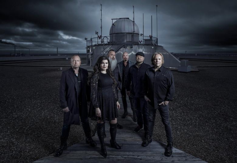 Kuvassa on keskellä Yö-yhtyeen viisi miestä ja Suvi Teräsniska keskellä kuvaa seisomassa. Heidän takanaan on suuri rakennus, jossa on pyöreä torni ja pystysuoria antenneja.