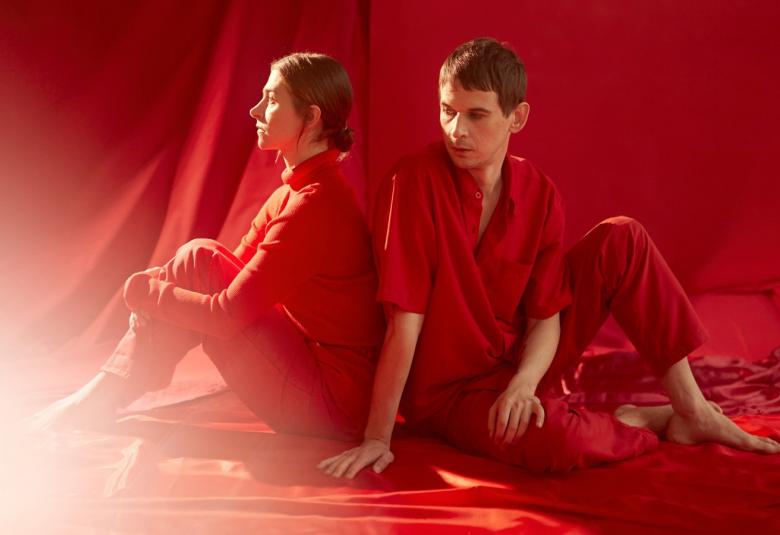 Kuvassa ovat nainen ja mies vinosti selät vastakkai.  He istuvat lattialla ja mies on kääntyneenä oikealle ja nojaa lattiaan toisella kädellään. Sekä vaatteet, lattia ja seinä ovat punaisia.  Vasemmasta kulmasta tulee valoa.