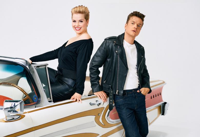 Kuvassa ovat valko-ruskea kuvioisen American raudan vieressä mustassa nahkatakissa ja farkuissa sekä avo-autossa istumassa mustassa mekossa Heta Halonen.  