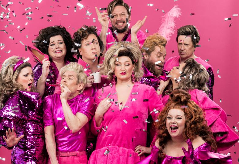 Kuvassa ovat näyttelijät tiiviisti keskellä olevan vaaleatukkaisen naisnäyttelijän ympärillä. Hänella on pinkki puki ja tausta on pinkki.