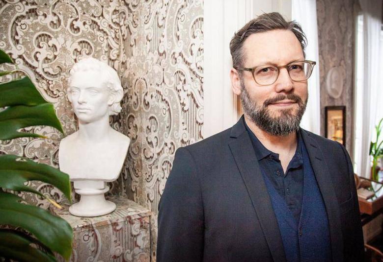 Kuvassa on Joel Haahtela seisomassa huoneessa, jossa on taustalla ruskehtava, koukeroinen tapetti.  Takana on myöös jalustalla valkoinen rintakuva patsas ja viherkasvin lehti.  Haahtelalla on tumman sininen paita päällä.