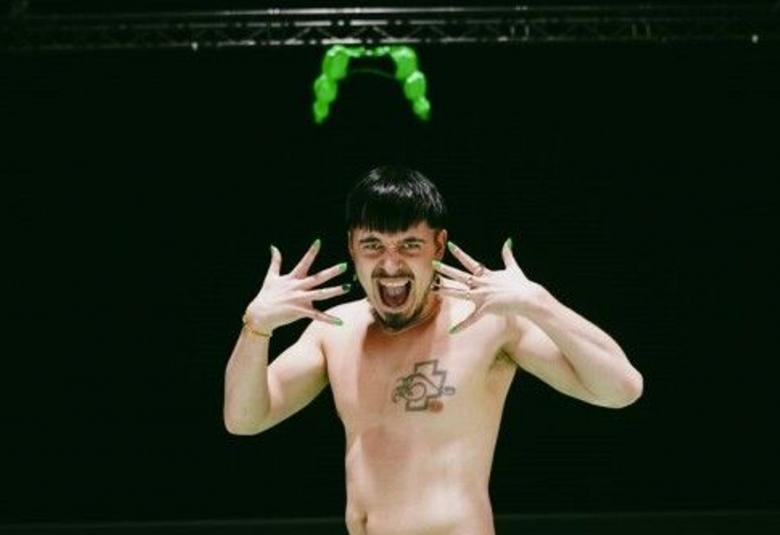 Kuvassa on Käärijä paidattomana ja rinnassaan näkyy Rammsteinin tatuointi.  Katossa on kirkkaan vihreä bolero.