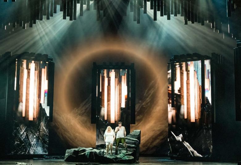 Kuvassa ovat kiven päällä valkoisissa puvuissa olevat nainen ja mies.  Taustalla on iso, ruskea pyöreä kuvio, jolla on kotkan siivet.  Taustalla on molemmin puolin turkooseja, ruskeita ja mustia valokennoja pystysuorassa.