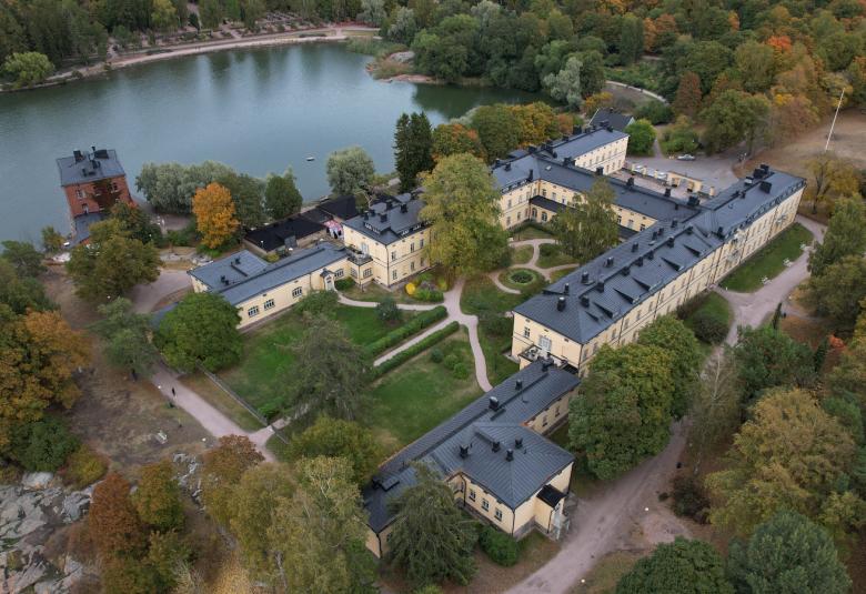 Kuvassa on Lapinlahden kellertävät sairaalarakenukset ja rannassa punatiilinen pieni rakennus. Rakennuksista kaksi on pitkiä ja ja niitä yhdistää lyhyempi rakennus keskellä. Taustalla näkyy vettä. Kuva on otettu ilmasta yläviistosta.