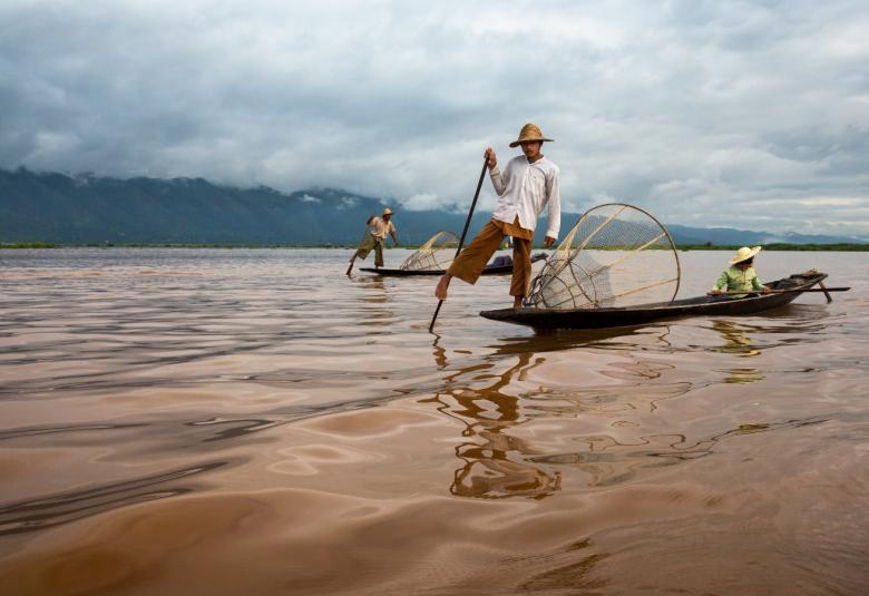  Kuvassa on kalastajia kahdessa veneessä seisomassa ja veneissä on kuljettaja. Taivas on pilvinen ja taustalla on metsää.  Vesi on ruskehtavaa.
