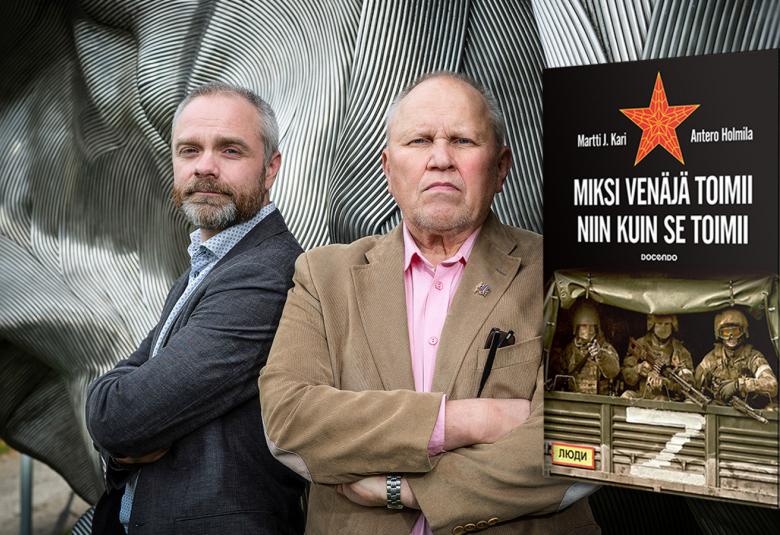 Kuvassa ovat Antero Holmila tumman harmaassa takissa ja Martti J. Kari vaalean ruskeassa takissa.  Miehet seisovat sivuttain.  Taustalla on harmaita kaaria.