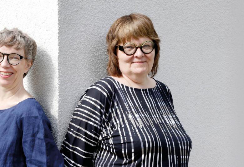 Kuvassa ovat Jaana Semeri sinisessä puserossa ja Leena Virtanen musta-valkoraidallisessa puserossa.  He nojaavat seinään ja välissä on vaalean seinän pieni kulma.