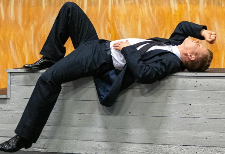 Kuvassa on mies mustassa puvussa makaamassa harmaan laatikon päällä ja taustalla on keltaista ruispeltoa.