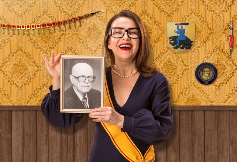 Kuvassa on Mia Vuorela kädessään Urho Kekkosen valokuva ja kelta-punaraitainen airutnauha. Taustalla on vaalean ruskeakuvioista tapettia ja alaosassa tummaa paneelilautaa.