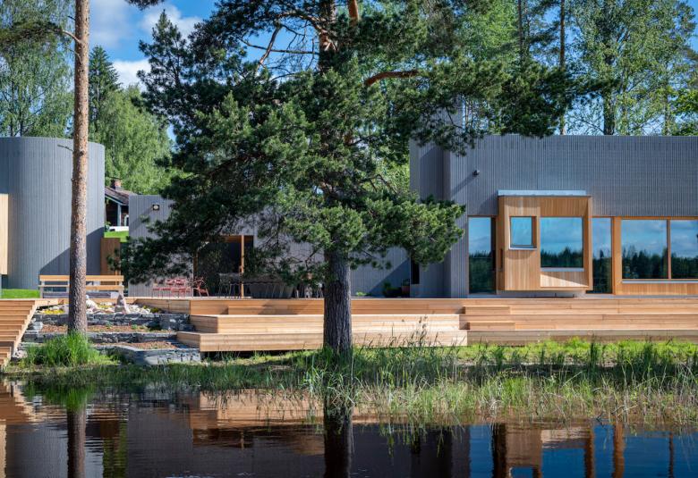 Kuvassa on Taidesaunan mustat rakennukset ja puun väriset laiturirakennelmat järven rannassa. Sauna on mäntyjen keskellä.