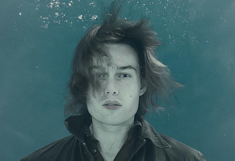 Kuvassa on hiukseet harallaan nuori mies, joka on puolivartalokuvassa ja mustassa paidassa. Hän on ikäänkuin turkoosissa vedessä ja yllä näkyy ilmakuplia.