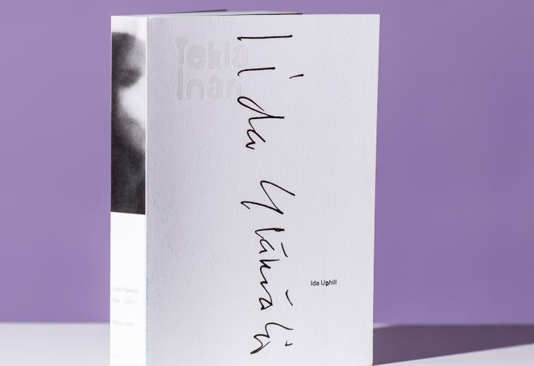 Kuvassa on vaalean violetit reunat ja keskellä on valkoista, jossa on Iida Ylämäen nimikirjoistus. Keskellä olevasta raosta näkyy harmaa hahmo.