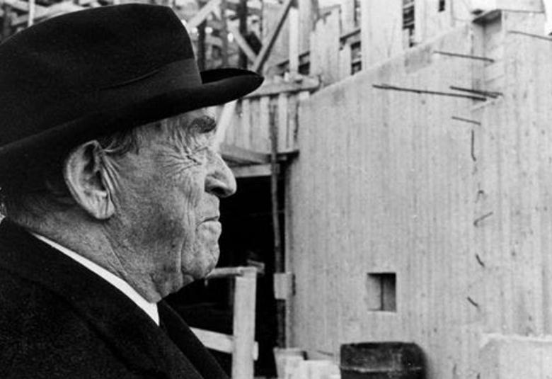 Kuvassa on vasemmalla edessä alvar Aalto katsomassa edessä olevaa rakennustyömaata, josta näkyy sementti seinää.  Kuva on musta-valkoinen.