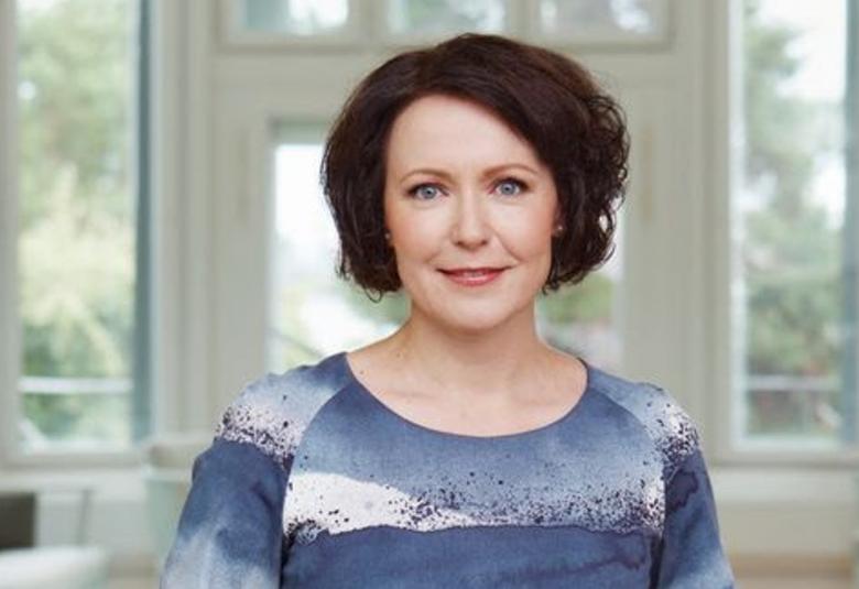 Kuvassa on Jenni Haukio seisomassa valkokarmisten ikkunoiden edessä. Hänellä on t-paita, jossa on vaalean sinisiä ja tumman sinisiä leveitä raitoja.