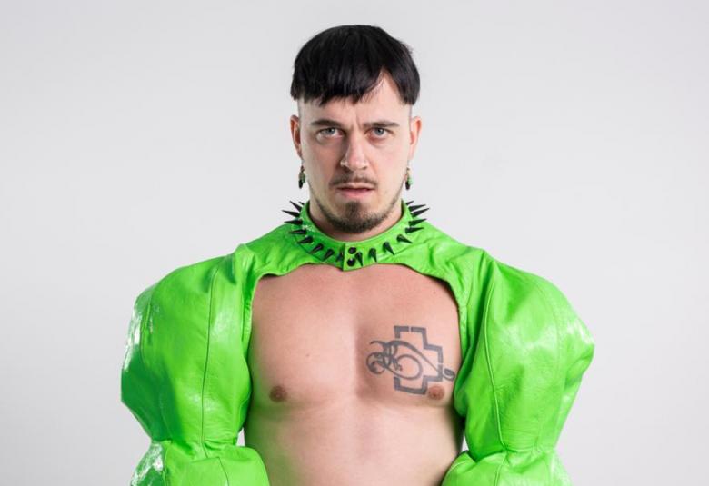 Kuvassa on Käärijä vaalean vihreässä bolerossaan puolivartalokuvassa.  Hänellä on rinta paljaana jaoikealla näkyy tatuointi ja kaulassa musta panta pystyniitteineen.