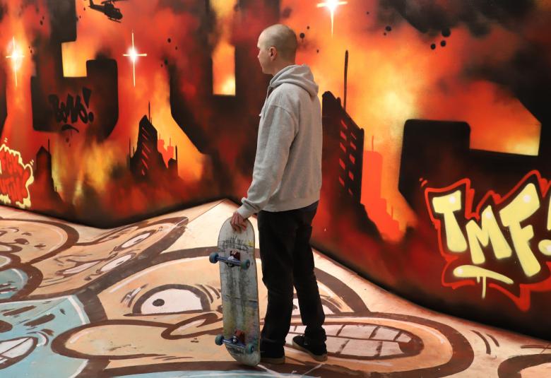 Kuvassa on aulatilassa värikkäitä graffitteja lattialla ja seinillä. Keskellä seisoo nuori mies skeittilauta kädessään. 