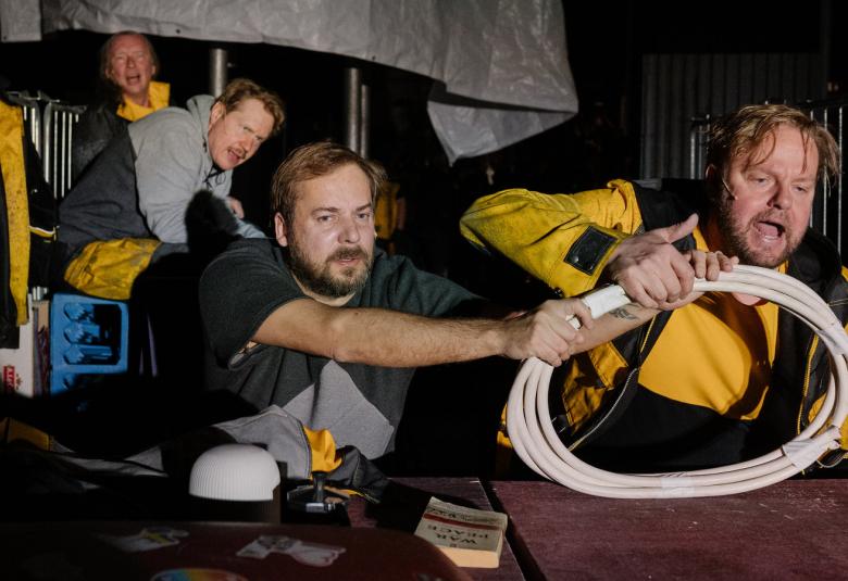 Kuvassa on kaksi miestä istumassa etualalla, joista toinen pitää valkoisesta letkukerästä kiinni oikealla kädellään ja toinen mies pitelee letkukerää myös.  Takana on kaksi miestä istumassa ja katselemassa. Miehillä on keltaisia takkeja ja mustat t-paidat.