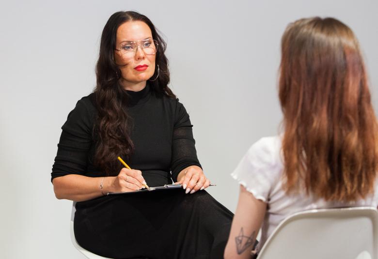 Kuvassa on Katriina Haikala istumassa ja piirtämässä mustassa puserossa. Vastapäätä on nainen valkoisessa puserossa.