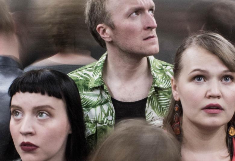 Kuvassa on alempana kahden nuorehkon naisen ja ylempänä heidän keskellään takana yhden miehen kasvot katse oikealle suunnattuna.  Takana on selin ihmisiä.