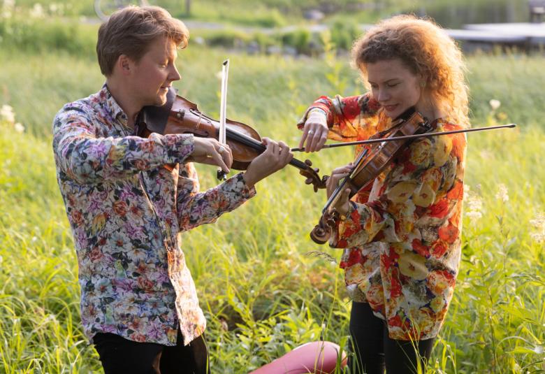 Kuvassa ovat Kuhmon Kamarimusiikin taiteelliset johtajat Antti Tikkanen ja Minna Pensola sivuttain soittamassa viulua.  He ovat keskellä heinikkoa vehreän kesäisessä maisemassa.