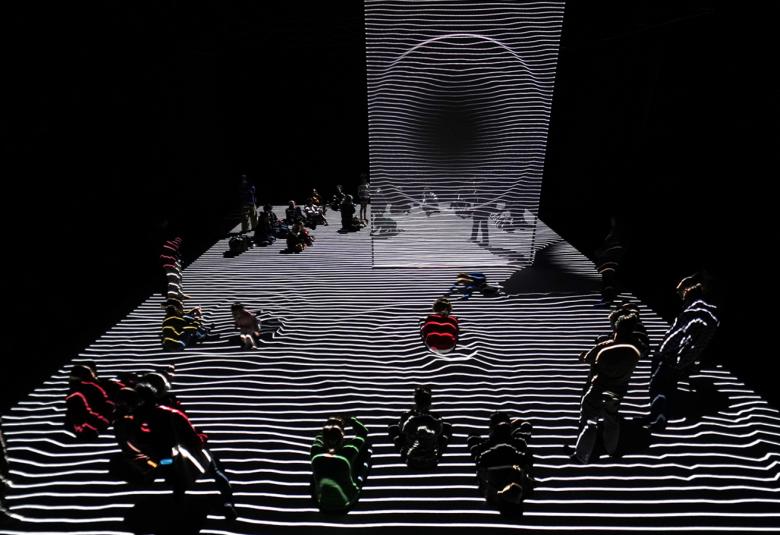 Kuvassa on mustalla pohjalla alaosassa vinosti ylöspäin valkoraitaista mattoa ja sen päällä on tummia ihmishahmojen ryhmiä.  Ylhäältä alas on valkoraitainen valkokangas.