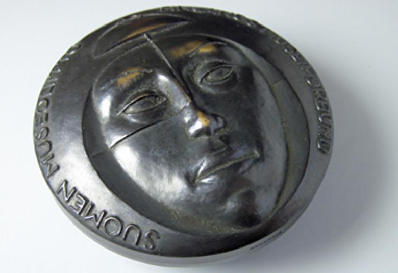 Kuvassa on tumma mitali, jossa on naisen kasvot.