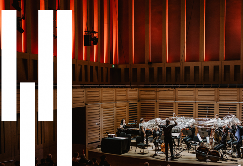 Kuvassa on orkesteri Riot Ensemble esiintymislavalla. Orkesterissa on alle kymmenen soittajaa ja heidän päällään on muovisuikaleita. Vasemmalla on valkoisia palkkeja pianomaisesti.