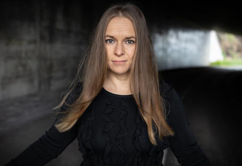 Kuvassa on Satu Rämö seisomassa taustallaan tumma tila, josta näkyy vasemmalta hieman valoa ja vihreää kiven raosta. Hänellä on pitkät ruskeat hiukset ja päällään musta t-paita.  