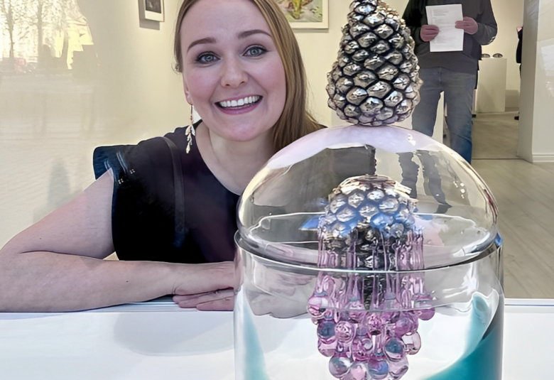 Kuvassa on Sini Majuri nojaa valkoiseen pöydän pintaan ja hänen edessään on lasiteos, jossa on hopeinen käpy ylöspäin ja se heijastuu alaspäin hopeisena käpynä sekä violettina alaspäin menevinä kävyn heijastumina.