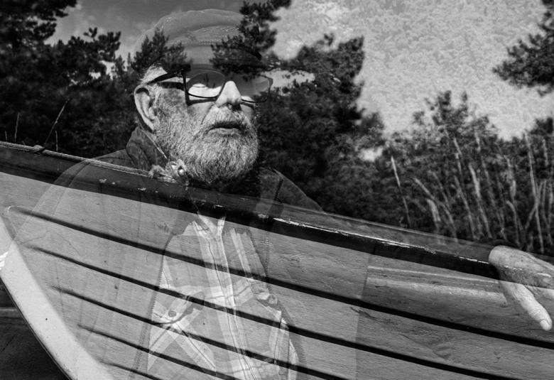 Kuvassa on mies puolivartalokuvassa ja hänen kätensä näkyy puuveneen päällä. Vene on läpinäkyvänä. Kuva on musta-valkoinen. 