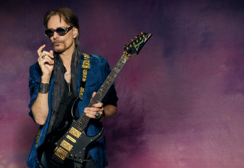 Kuvassa on Steve Vai kitara kädessään. Hänellä on tumman sininen paita päällään.  Tausta on tumman violetti ja siinä on höyryä.