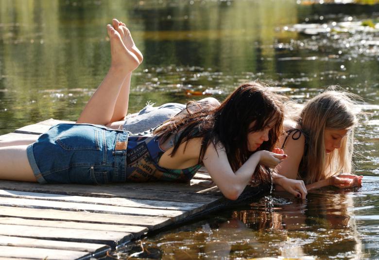 Kuvassa on kaksi tyttöä laiturilla makaamassa ja katsomassa alas rantaveteen, joka on vihertävän sinistä.  