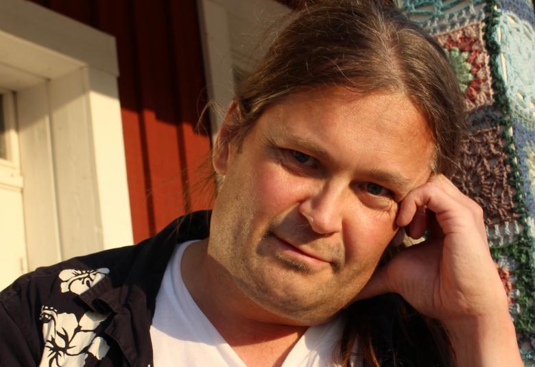 Kuvassa on Jani Saxellin kasvokuva ja hänen takanaaan näkyy punaista taloa ja valkoisia ikkunapuitteita.