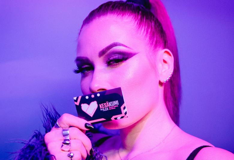 Kuvassa on BESSin kasvokuva ja hänellä on huuliensa päällä ja oikeassa kädessään kesäkuvipaketti.  BESSillä on poninhäntä. Kuvan sävy on lila koko kuva-alalla.