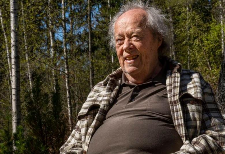 Kuvassa on Jorma Panula istumassa ja hänellä on ruskea pusero ja sen päällä vaalearuudullinen flanellipaita. Ympäristö on vihreä.