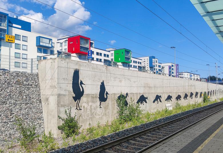 Kuvassa menee etualalla junaraide ja oikeassa sivussa on katosta.  Vasemmalla ylhäällä näkyy valkoista kerrostaloriviä. Siinä on ulokkeina sinisiä, punaisia ja vihreitä ulokeparvekkeita. Radan vieressä on betoniseinämää ja siinä on mustan lennosta laskeutuvan kurjen kuvasarjaa pitkin seinämää.