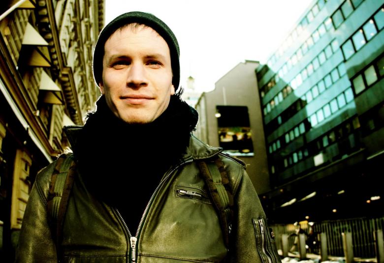 Kuvassa on Akse Pettersson taustallaan kaupunkirakennuksia molemmin puolin.  Hänellä on musta pipo päässään.  Kuva on puolivartalokuva.