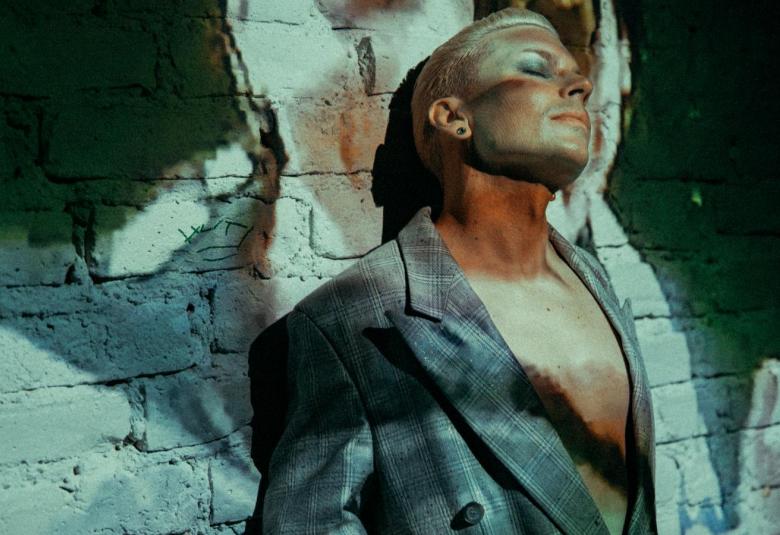 Kuvassa on Antti Tuisku nojaa tiiliseinään, josta osa on tummaa varjoa ja osa valkoista erityisesti Tuiskun takana.  Tuisku katsoo oikealle ja ottaa aurinkoa.  Hänellä on tummahko takki päällään ilman aluspaitaa.