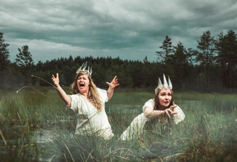 Kuvassa ovat Marja Hepo-aho ja Sini Majuri ovat vyötäröään myöten suossa ja ojentavat käsiään viistosti ylöspäin.  Heillä on muinaispuvut päällään ja lasikruunut päässään.  Taustalla on harmaata pilveä.