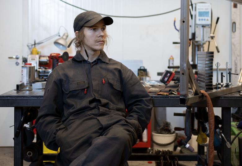 Kuvassa on Eetu Huhtala istumassa työpöytänsä edessä.  Pöytä on täynnä erilaisia  työkaluja ja taustalla näkyy valkoista kuperaa seinää.  