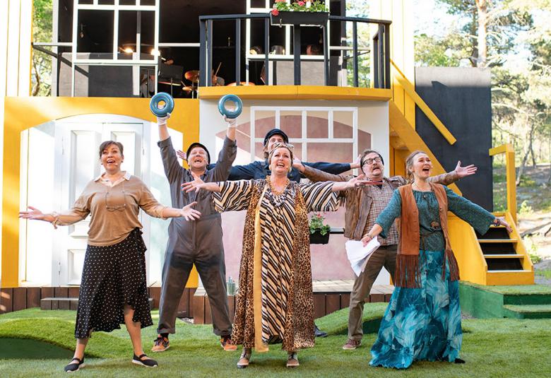 Kuvassa on kolme naista eturivissä kesämekoissa ja takana kolme miestä.  Kaikki laulavat ja ovat kädet ylhäällä ojennettuina.  Taustalla on talomaista kulissia ja taustalla vihreitä puita.